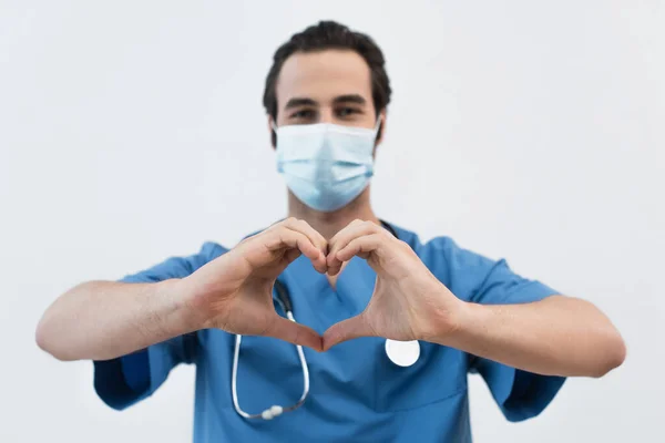 Medico sfocato in maschera medica che mostra il simbolo del cuore con le mani isolate sul grigio — Foto stock