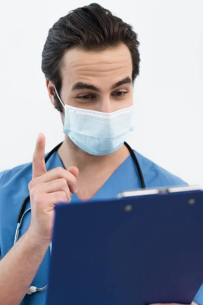 Врач в медицинской маске показывает жест идеи рядом с размытым блокнотом, изолированным на сером — стоковое фото