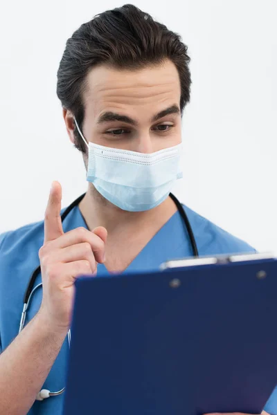 Doctor in medical mask showing idea gesture near blurred clipboard isolated on grey — Stock Photo