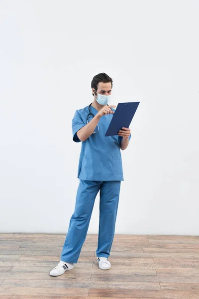 Full length view of doctor in medical mask pointing at clipboard near grey wall — Stock Photo