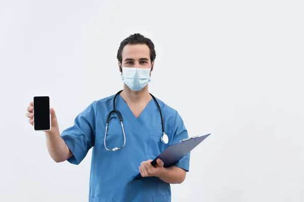 Arzt in medizinischer Maske hält Klemmbrett und Smartphone mit leerem Bildschirm isoliert auf grau — Stockfoto