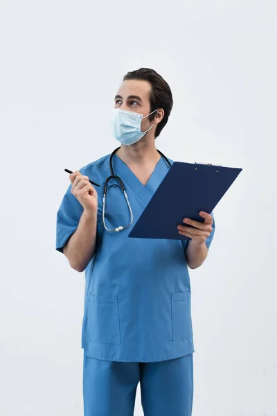 Врач в медицинской маске смотрит в сторону и указывает ручкой, изолированной на сером — стоковое фото