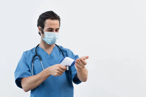 Молодой врач в медицинской маске, измеряющий собственную температуру пирометром, изолированным на сером — стоковое фото