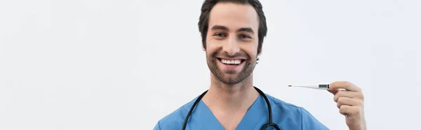 Веселый врач держит электронный термометр, улыбаясь в камеру, изолированную на сером, баннер — стоковое фото