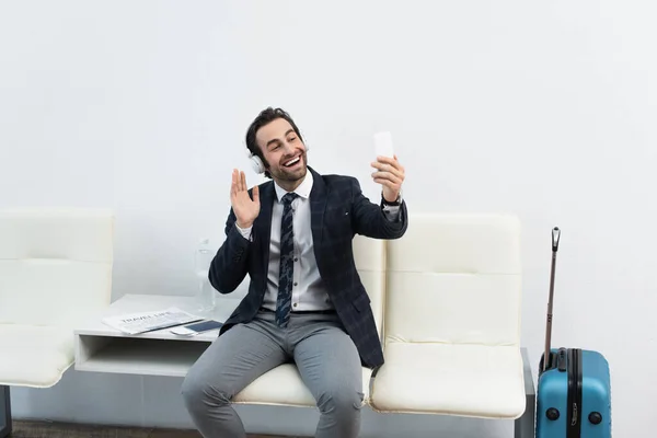 Hombre alegre en auriculares saludando de la mano mientras toma selfie en el teléfono inteligente en la sala de salida - foto de stock