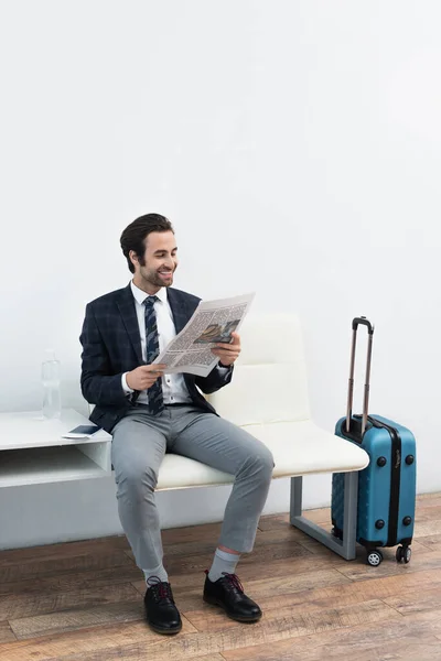 Sonriente hombre leyendo periódico cerca de bolsa de viaje en la sala de salida - foto de stock