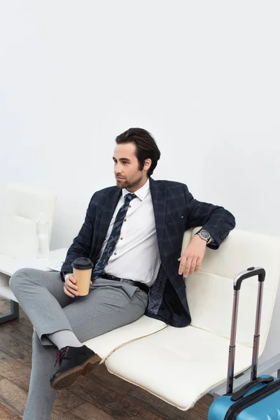 Joven sosteniendo taza de papel mientras está sentado en la sala de salida cerca de la bolsa de viaje — Stock Photo