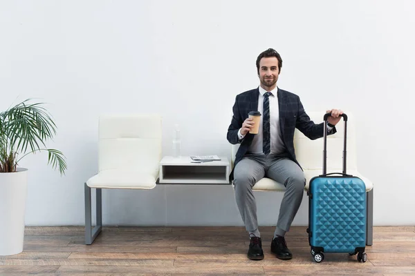 Tourist mit Pappbecher lächelt in die Kamera, während er in der Abflughalle neben dem Koffer sitzt — Stockfoto