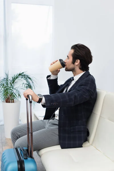 Traveler sitting in departure lounge near suitcase and drinking coffee from paper cup — Stock Photo