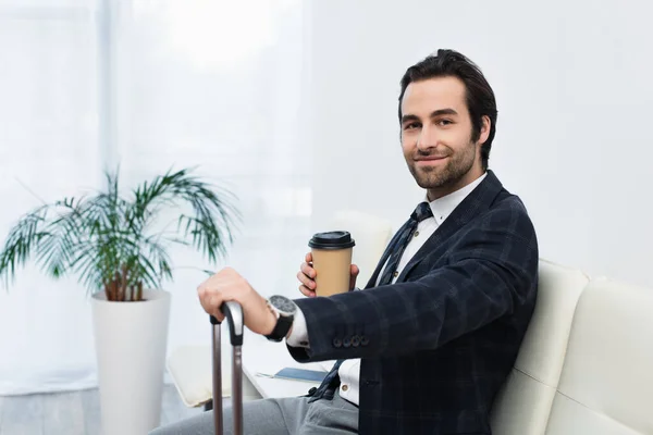 Heureux voyageur avec café pour aller sourire à la caméra en attendant dans le salon de départ — Photo de stock