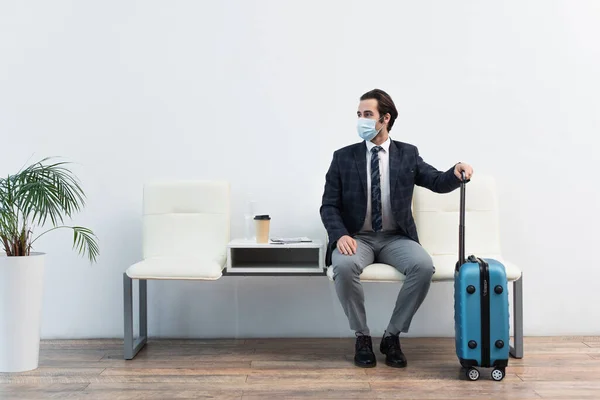 Мандрівник у медичній масці, сидячи у вітальні відправлення поблизу валізи та дивлячись подалі — стокове фото