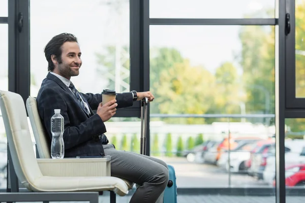 Glücklicher Mann sitzt mit Pappbecher und Koffer in der Abflughalle — Stockfoto