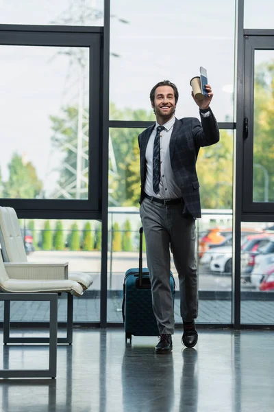 Hombre feliz con maleta, taza de papel y pasaporte mirando hacia otro lado mientras camina en la sala de salida - foto de stock