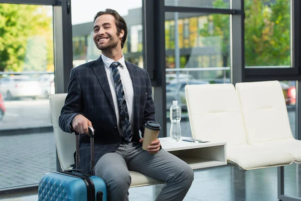 Uomo sorridente che distoglie lo sguardo mentre è seduto nella sala partenze con bicchiere di carta e valigia — Foto stock