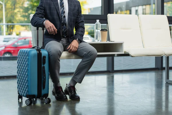 Vue recadrée du voyageur assis dans le salon de départ près de la valise, bouteille d'eau et tasse en papier — Photo de stock