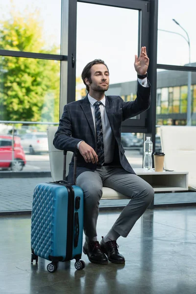 Hombre mirando hacia otro lado y saludando la mano mientras espera en la sala de salida cerca de la bolsa de viaje - foto de stock