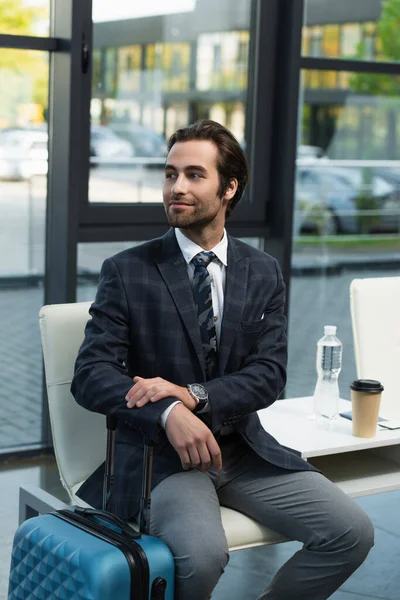 Positive man looking away in departure lounge near suitcase, coffee to go and bottle of water — Stock Photo