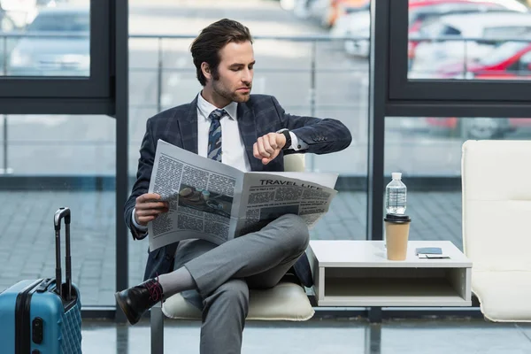 Man with travel life newspaper looking at wristwatch while sitting in departure lounge — Stock Photo