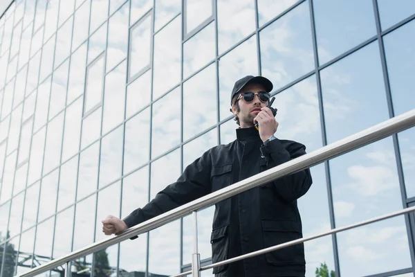 Vue à angle bas de la garde en uniforme noir et lunettes de soleil parlant sur talkie-walkie près du bâtiment avec façade en verre — Photo de stock