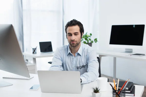 Junger Sicherheitsmann arbeitet an Laptop in der Nähe verschwommener Monitore im Überwachungsraum — Stock Photo