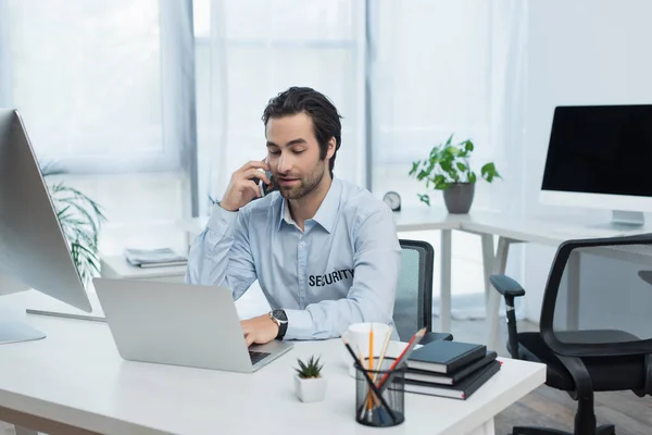 Позитивный человек безопасности с помощью ноутбука во время разговора на смартфоне в офисе — стоковое фото