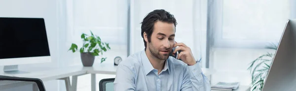 Hombre de negocios positivo hablando en el teléfono móvil cerca de monitores de ordenador en la oficina, bandera - foto de stock