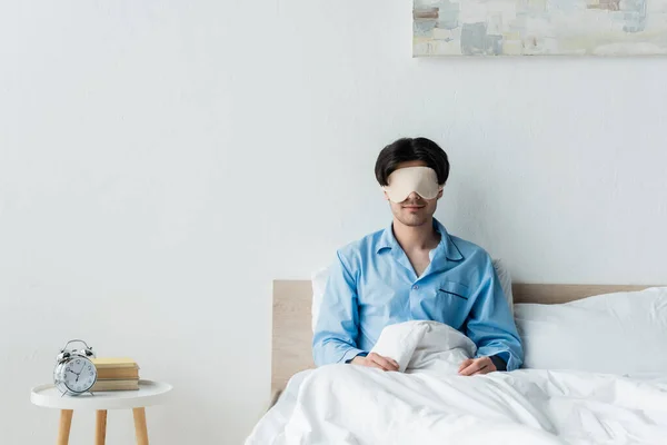 Joven morena hombre sentado en la cama en pijama azul y la máscara del sueño - foto de stock