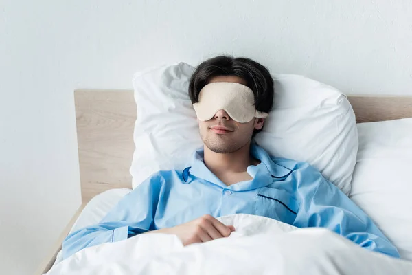 Brunetta uomo in pigiama blu e maschera del sonno sdraiato su biancheria da letto bianca — Foto stock