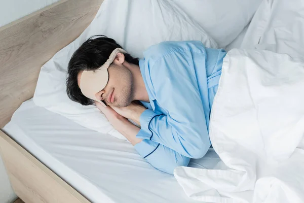 Joven con máscara para dormir y pijama durmiendo sobre ropa de cama blanca - foto de stock