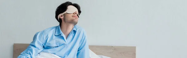 Jeune homme en masque de sommeil et pyjama bleu assis sur le lit le matin, bannière — Photo de stock