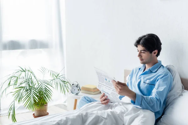 Young man in blue pajamas reading newspaper near vintage alarm clock on bedside table — Stock Photo