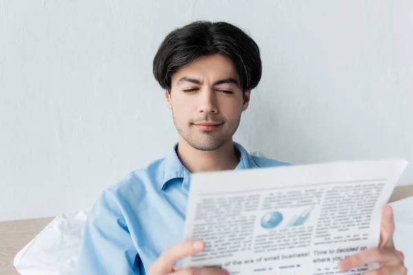 Hombre joven y positivo en pijama azul leyendo el periódico en el dormitorio - foto de stock