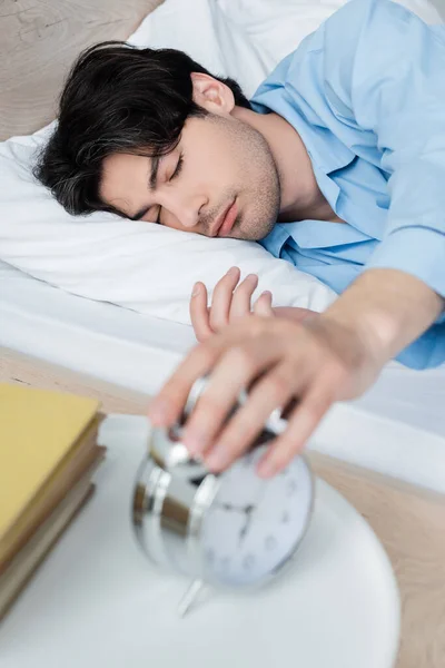 Man with closed eyes lying in bed and turning off blurred alarm clock — Stock Photo