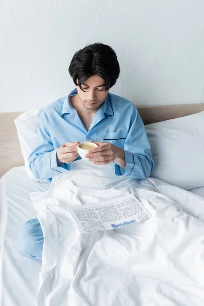 Brunette homme en pyjama bleu tenant tasse de café près du journal sur literie blanche — Photo de stock