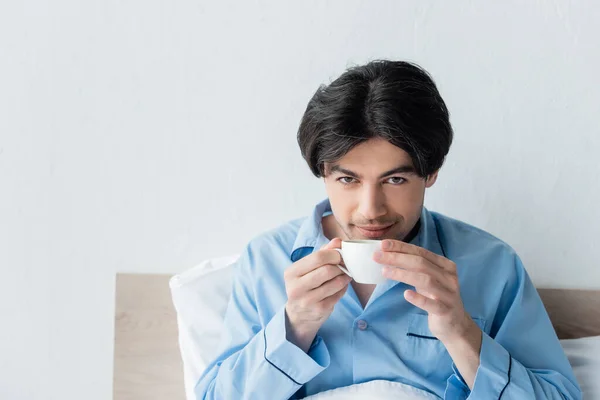 Bruna uomo sorridente a macchina fotografica mentre beve caffè in camera da letto — Foto stock