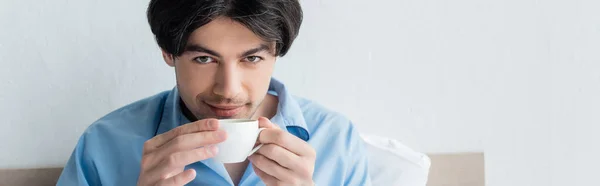 Jeune homme brune souriant à la caméra tout en buvant le café du matin, bannière — Photo de stock