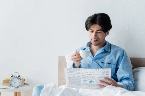 Hombre positivo leyendo el periódico de la mañana y beber café en el dormitorio - foto de stock