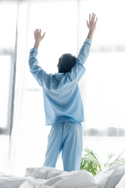 Vista trasera del hombre en pijama azul estirándose con las manos levantadas cerca de la ventana - foto de stock