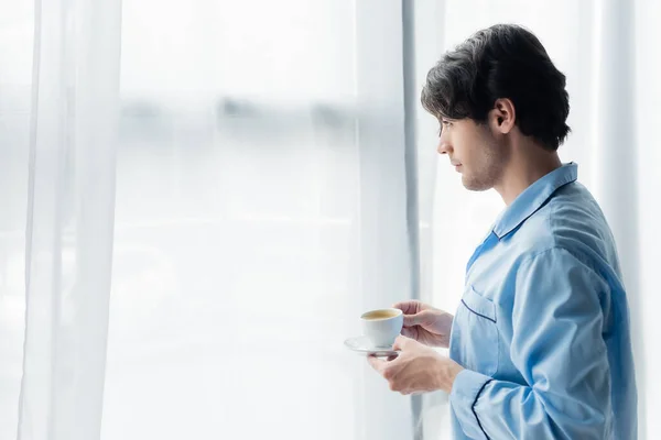 Vue latérale de brune homme en pyjama bleu debout avec tasse de café près de la fenêtre — Photo de stock