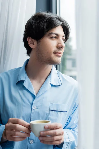 Junger brünetter Mann im blauen Pyjama steht mit Kaffeetasse am Fenster — Stockfoto