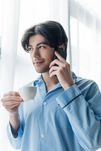 Uomo sorridente in pigiama blu con in mano una tazza di caffè mentre parla sul cellulare — Foto stock