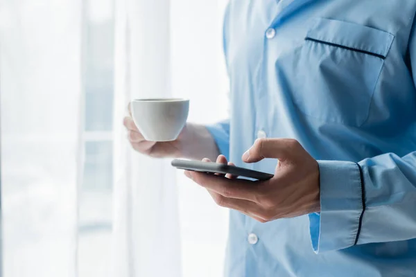 Vista parziale dell'uomo in pigiama blu con smartphone e tazza di caffè — Foto stock