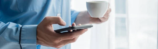 Vista recortada del hombre en pijama azul usando teléfono celular mientras sostiene la taza de café, pancarta - foto de stock