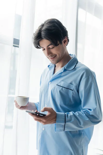 Uomo positivo in pigiama blu che tiene la tazza di caffè e chatta sullo smartphone vicino alla finestra — Foto stock