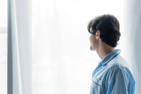 Jeune homme brune en pyjama bleu debout près de la fenêtre avec rideau blanc à la maison — Photo de stock