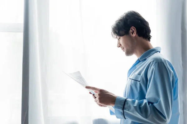 Vista lateral del joven sonriente en pijama azul leyendo el periódico matutino cerca de la ventana - foto de stock