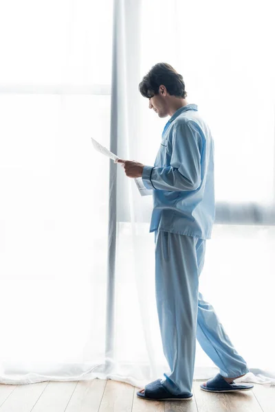 Full length view of young man in blue pajamas reading newspaper near window — Stock Photo