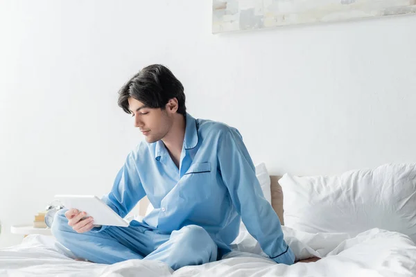 Молодой человек в синей пижаме смотрит на цифровой планшет, сидя на кровати со скрещенными ногами — стоковое фото