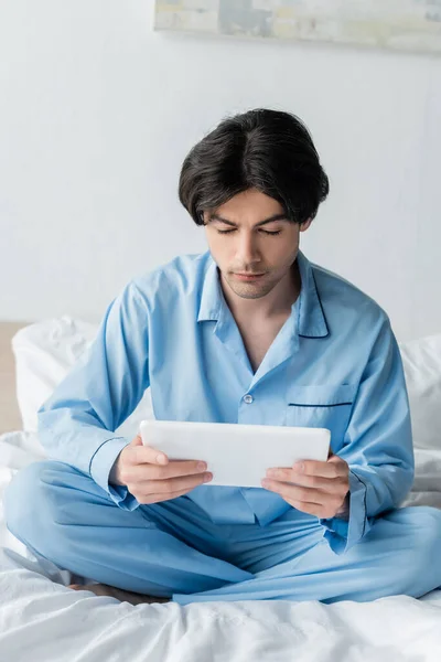 Brünetter Mann mit digitalem Tablet sitzt mit gekreuzten Beinen auf weißem Bettzeug — Stockfoto