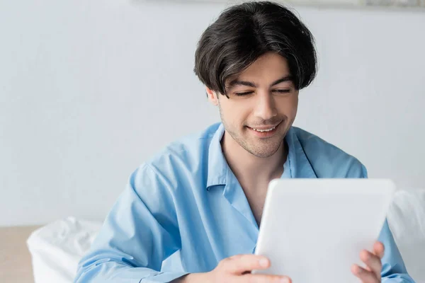 Sorrindo homem de pijama azul usando tablet digital no quarto — Fotografia de Stock